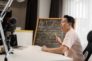 Instructor lectures at a camera and points to a chalk board