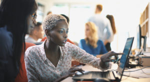Closeup side view of two mid 20's black women doing their design project on a computer. They might be software developers as well. Blurry coworkers in background, all released.