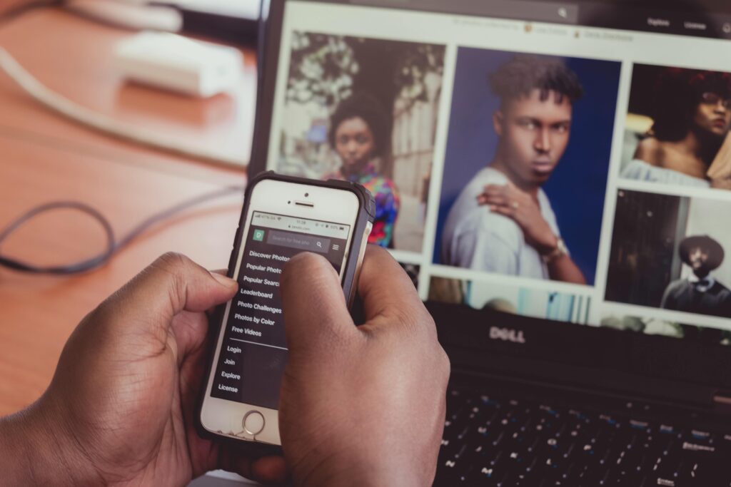 Close-up of hands holding smartphone in front of a laptop displaying images, indoors.