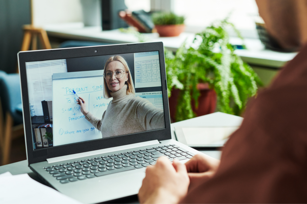Laptop screen with mature blond confident teacher in eyeglasses pointing at whiteboard while explaining Present Simple tense to online student