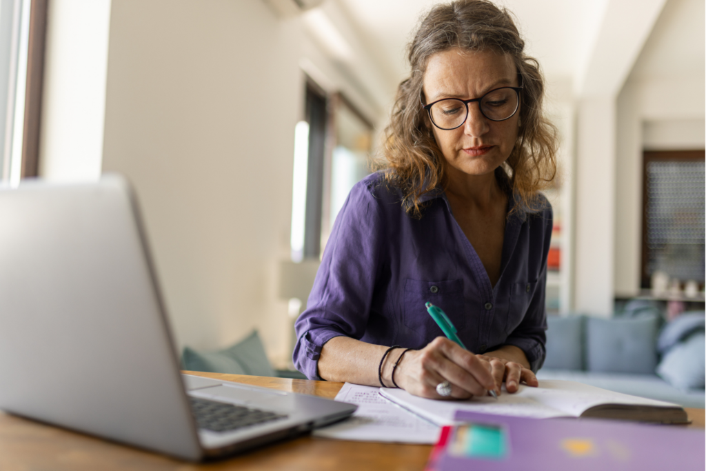 Beautiful mature adult woman, may be a professor or business woman, sits at kitchen counter with laptop, notebook and documents. She is modern professional woman dressed in smart casual clothes.