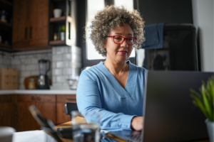 A mature woman with curly gray hair and glasses works on a laptop in a cozy home kitchen. The setting is relaxed, with natural light coming in through the window, creating a productive atmosphere