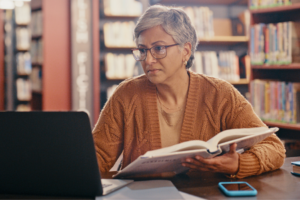 Senior woman, computer and planning in a library with book and laptop for lecture class. Teacher, university professor and education worker reading web data and research for class on a study desk
