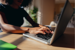 Close-up view of female hands typing on a laptop keyboard, working remotely in a home office, with sunlight streaming onto a wooden table, creating a warm and inviting atmosphere