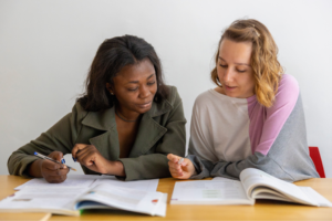 Two diverse women sitting at a table studying, reading books, and writing notes, focusing on educational concepts and language instruction in a school environment.