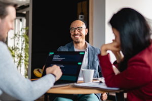Middle aged colleagues sitting around a table and discussing ideas while viewing an AI chatbot interface on a laptop during a collaborative business meeting in a modern office.