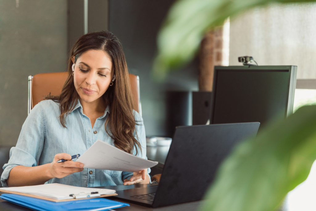 A mid adult businesswoman reviews documents while working in her office. An open laptop as well as a desktop pc are on her desk.