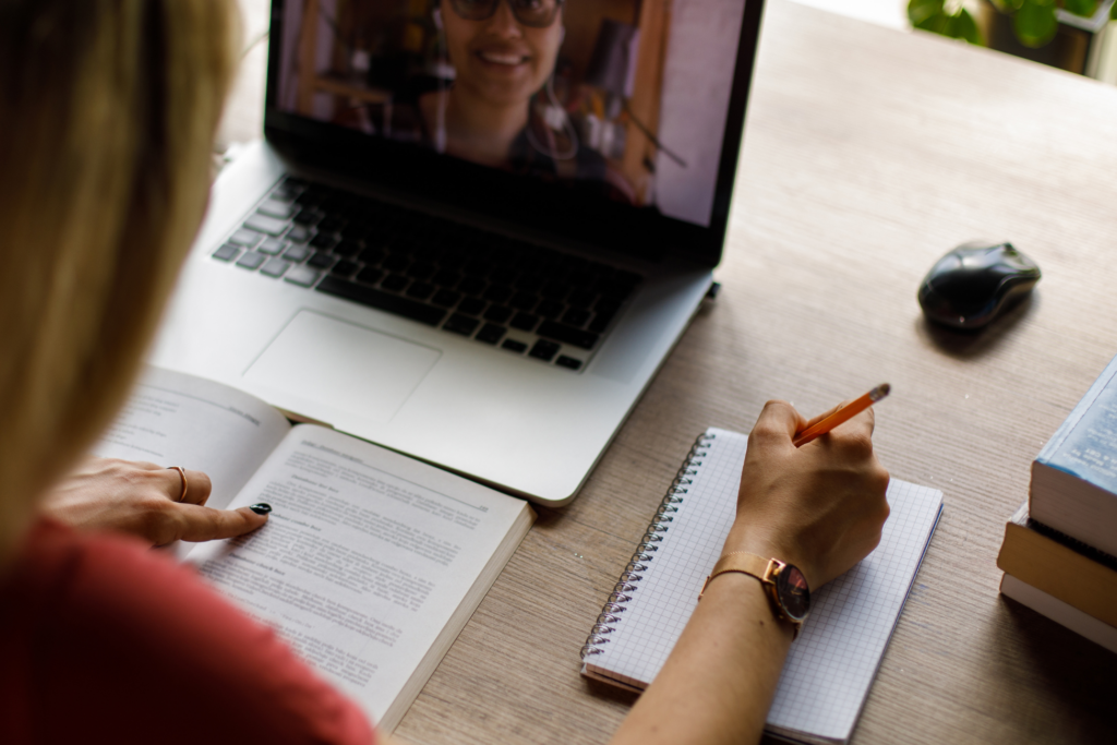 Over the shoulder view of young female university student sitting at her desk, reading a text in a book in front of her and writing notes while her professor is teaching an online class via video call that she is attending.