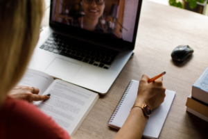 Over the shoulder view of young female university student sitting at her desk, reading a text in a book in front of her and writing notes while her professor is teaching an online class via video call that she is attending.