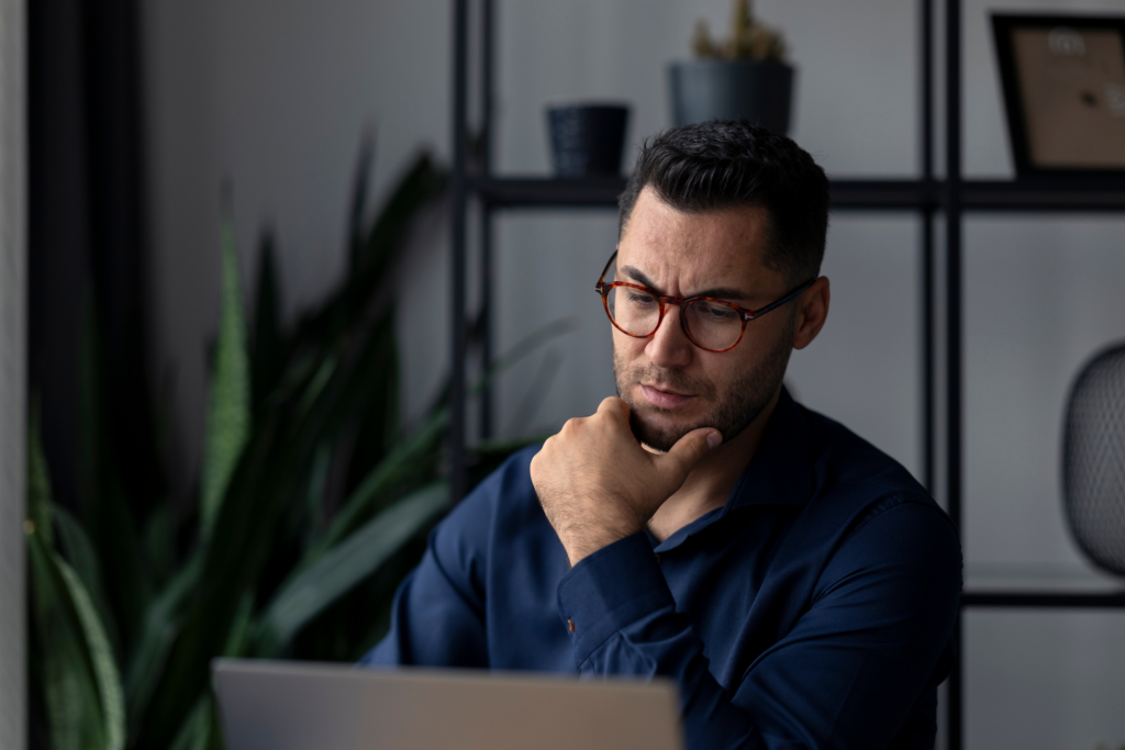 Focused young adult businessman working from home, using a laptop while pondering solutions with his hand resting thoughtfully on his chin, navigating challenges of remote work