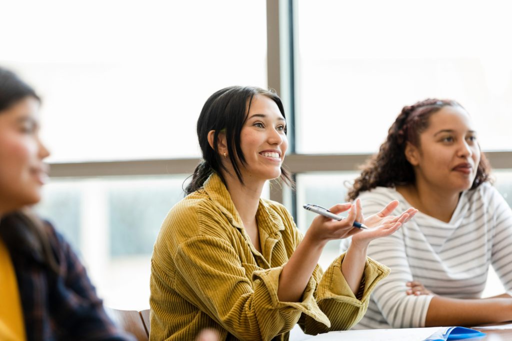 A young adult female college student smiles and gestures as she asks the unseen professor a question.