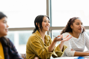 A young adult female college student smiles and gestures as she asks the unseen professor a question.