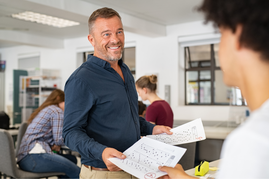 Happy mature professor giving corrected answer sheets to student in class. Rear view of black guy receiving exam test result in college classroom. Smiling proud teacher man returning test with marks to students in high school class.