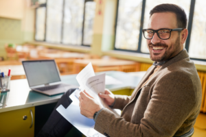 Happy male professor going through educational exam in the classroom and looking at camera.