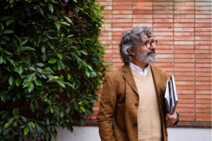 Mature professional man stands outdoors by a brick wall holding a laptop and documents, looking thoughtful.