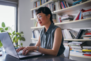 A wide angle view of a smiling Japanese entrepreneur typing on her laptop while sitting at the office desk.