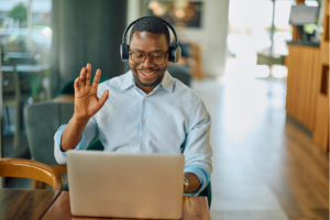 Young adult businessman wearing headphones, smiling and waving while engaging in a video call on his laptop at a cozy cafe