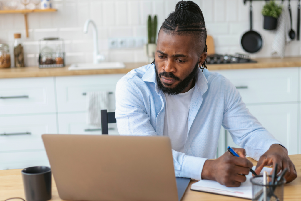 Focused African American man uses laptop computer for online learning remotely at home taking notes on notepad during online lesson