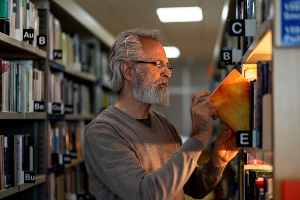 Close-up side shot with blurred background of a senior Caucasian retired man picking up a book of his interest at the city public library during the afternoon.