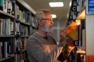 Close-up side shot with blurred background of a senior Caucasian retired man picking up a book of his interest at the city public library during the afternoon.