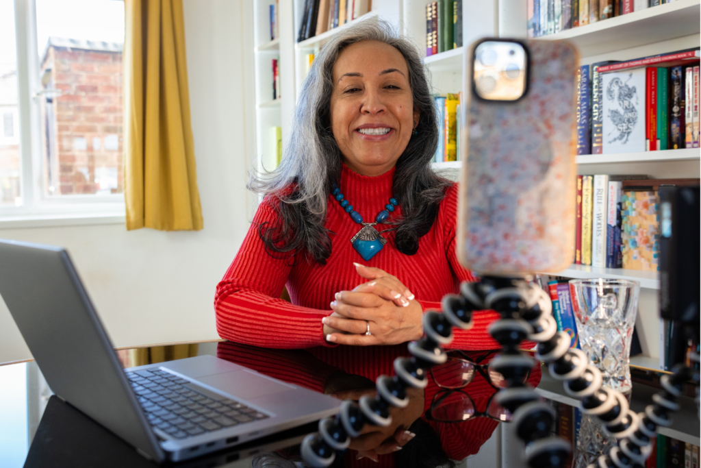 Medium shot of a mature woman sitting down at her dining table in her home in the North East of England. She has her laptop open and is using her mobile phone on a tripod to record herself as she is an influencer. The woman is looking at her phone while talking and smiling. A small light is shining on her to complete the set-up.
