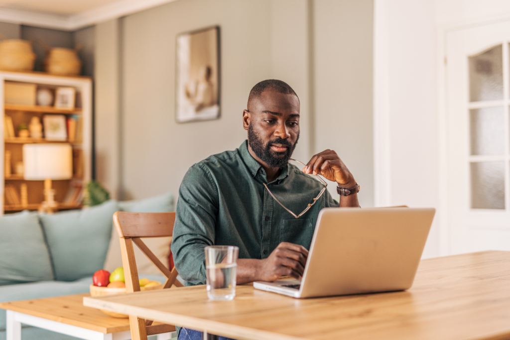 Concentrated young adult black man working from home, sitting at wooden table, using laptop, holding eyeglasses in hand, glass of water nearby