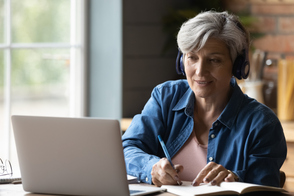 60s woman sit at table wear headphones take notes gain new skills use laptop and internet resources, on-line services. Video call event, modern tech, counselling, receive information remotely concept