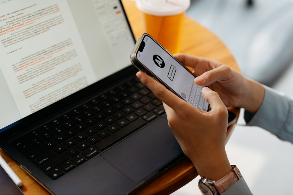 Asian woman using a smartphone with an AI assistant while seated at a desk with a laptop displaying a document. The integration of artificial intelligence into daily business and communication tasks.