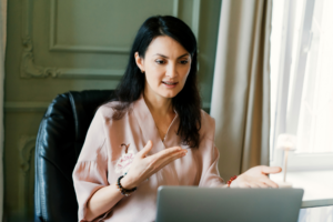 Professional woman speaking to a laptop during a video call about healthy longevity and wellness, using expressive hand gestures in a comfortable home office setting.