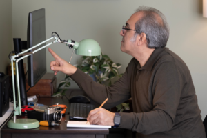 Man points at his computer screen while writing in a notebook