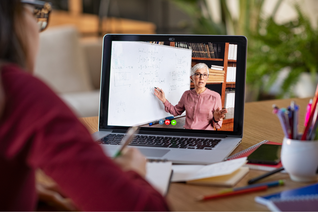 Young woman taking notes while looking at computer screen following professor doing math on video call.