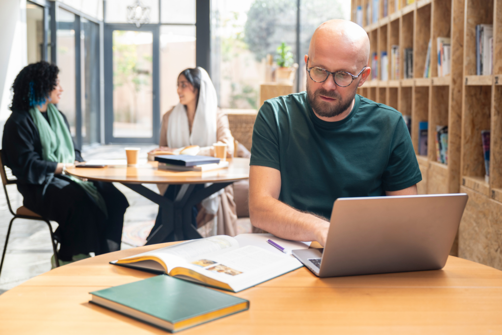 Bearded individual wearing eyeglasses and casual t-shirt sitting at round table, looking at open book, typing. Female students talking at table in background.