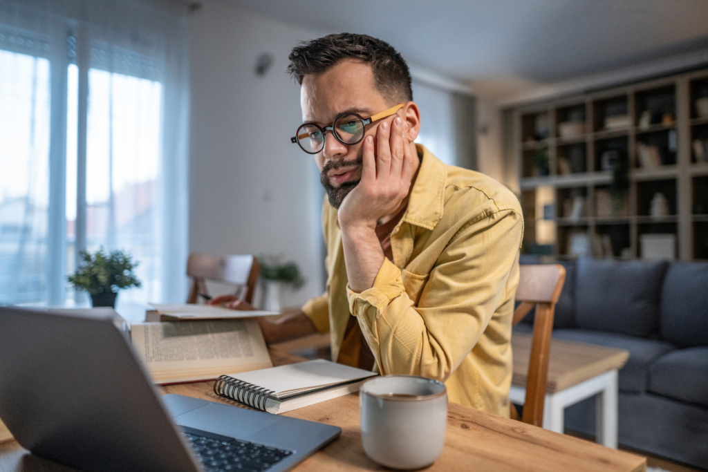 Adult man in glasses concentrating on work from home, sitting at a wooden table with a laptop, books, and coffee cup, actively engaging in remote study or telework