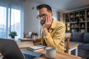 Adult man in glasses concentrating on work from home, sitting at a wooden table with a laptop, books, and coffee cup, actively engaging in remote study or telework