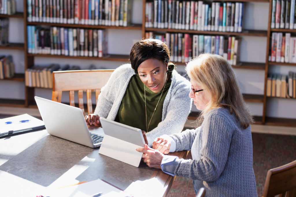 wo multi-ethnic women working together in the library. They are sitting at a table, one with a laptop and the other using a digital tablet, sharing information. The young African-American woman is in her 20s and her friend is a senior woman in her 70s. Perhaps they are students in a continuing education class working on a project.
