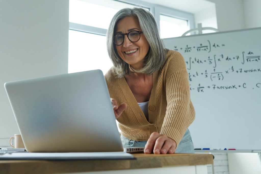 Mature woman teaching mathematics while standing near the whiteboard and looking at laptop