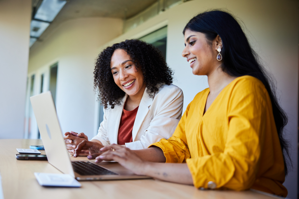 Two diverse young businesswoman talking and working on a laptop together at a desk in an office lounge area