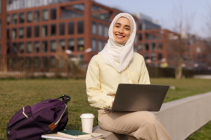 Student sits on laptop computer and smiles at the camera on a college campus