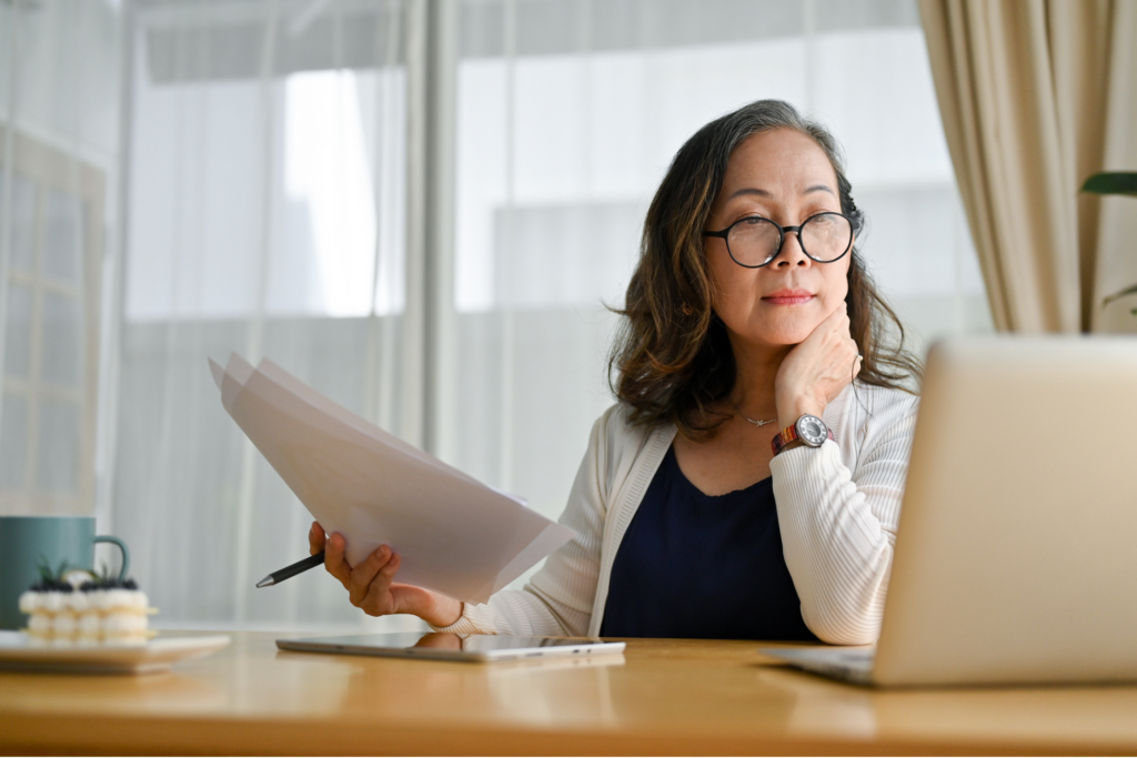 Concentrated asian middle aged female teacher or businesswoman in glasses sitting at desk using portable computer and examining paperwork.