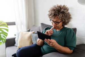 A middle-aged woman with curly hair, glasses, and a green sweater sits by a large window, smiling while reading an e-book on her reader, enjoying a relaxing moment at home.