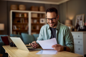 A smiling male college professor reading a student's essay paper in his office in front of a laptop.
