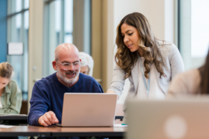 Teacher assists mature adult man with laptop computer in classroom